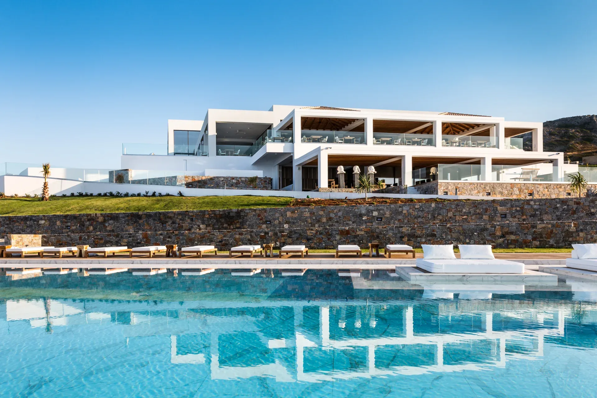 Modernes, weißes Luxusresort (Abaton Island Resort & Spa) auf Kreta mit großer Terrasse und Infinity-Pool im Vordergrund; klare Spiegelung des Gebäudes im türkisfarbenen Wasser unter strahlend blauem Himmel.