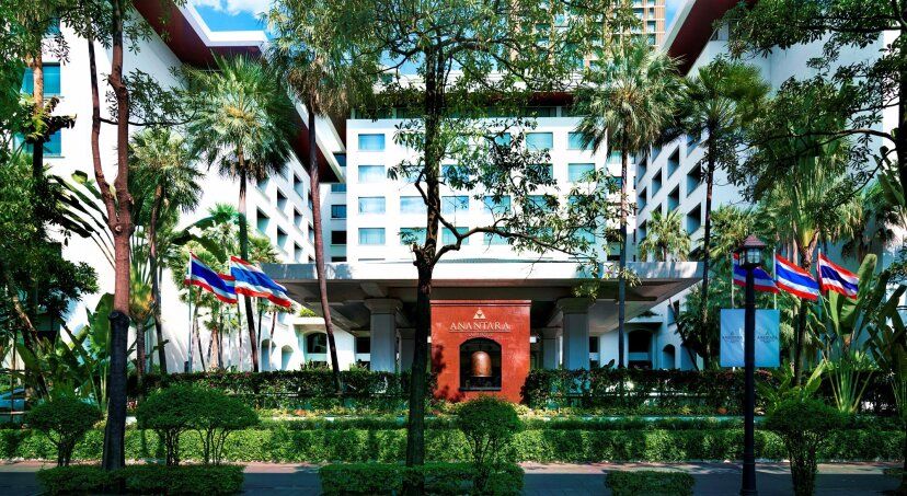 Elegante Eingangsfassade des Anantara Siam Bangkok Hotels, umgeben von üppigem Grün und tropischen Palmen, mit thailändischen Flaggen und moderner Architektur im Hintergrund.