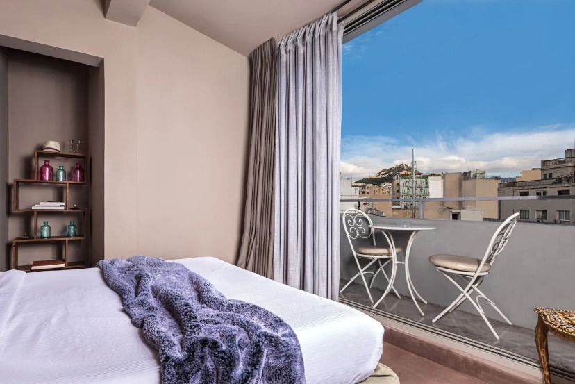 Modernes Zimmer im Andronis Athens mit Balkon und Stadtblick auf den Lykabettus-Hügel, ausgestattet mit gemütlichem Bett, minimalistischer Deko und eleganter Sitzgelegenheit im Freien.