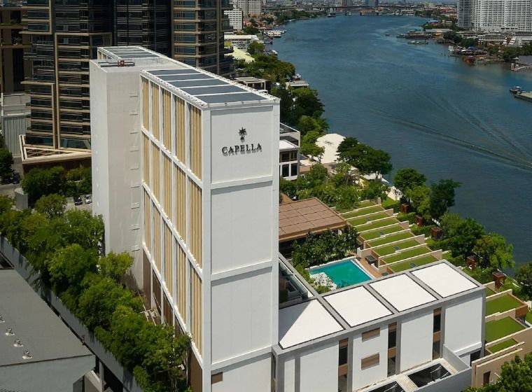 uxushotel Capella Bangkok am Ufer des Chao Phraya mit moderner weißer Fassade, Poolbereich und begrünten Terrassen, umgeben von Bäumen und mit Blick auf den Fluss und die Skyline.
