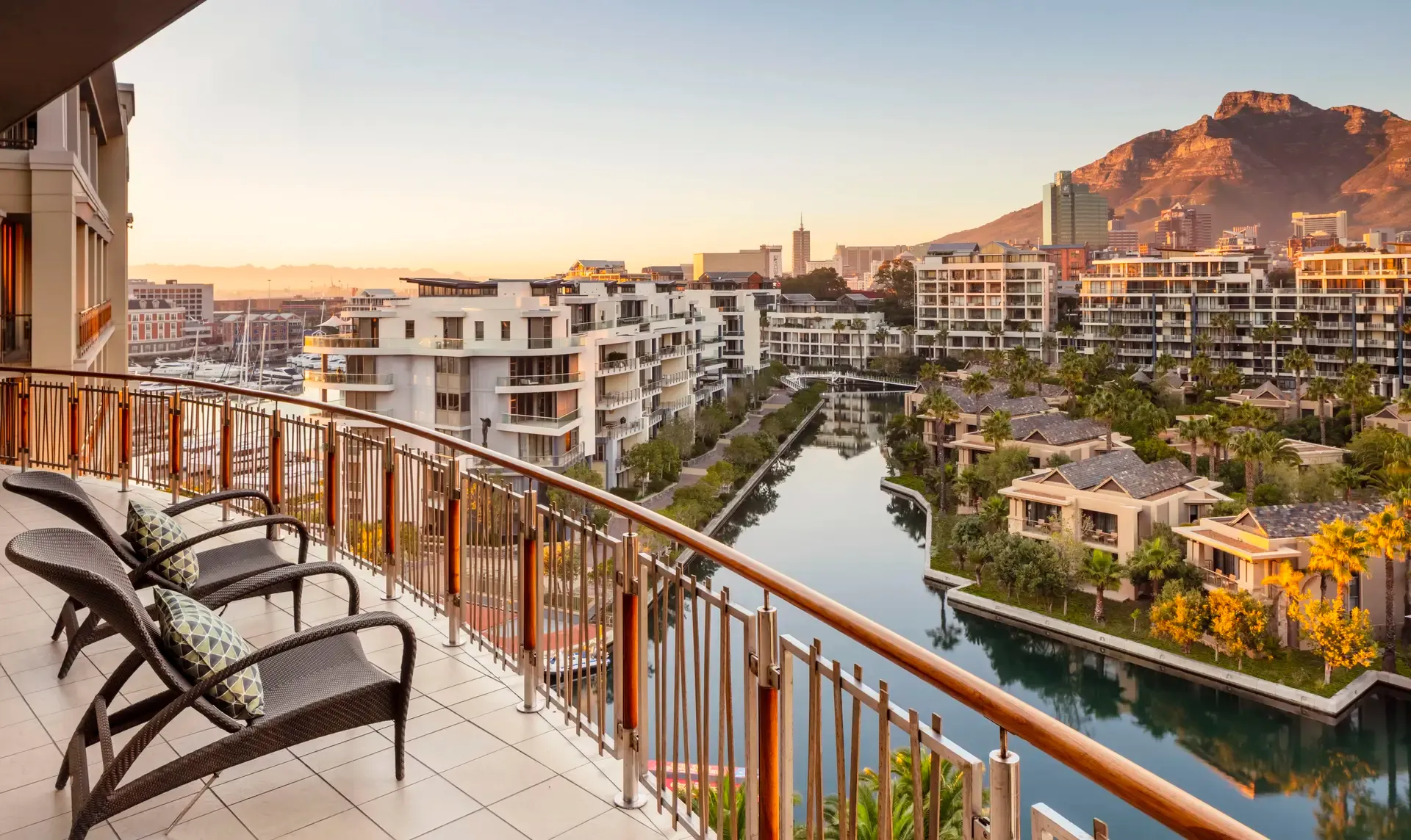 Aussicht vom Balkon des One&Only Cape Town mit Blick auf moderne Wohnanlagen, eine ruhige Wasserstraße und den Tafelberg im Hintergrund bei Sonnenuntergang.
