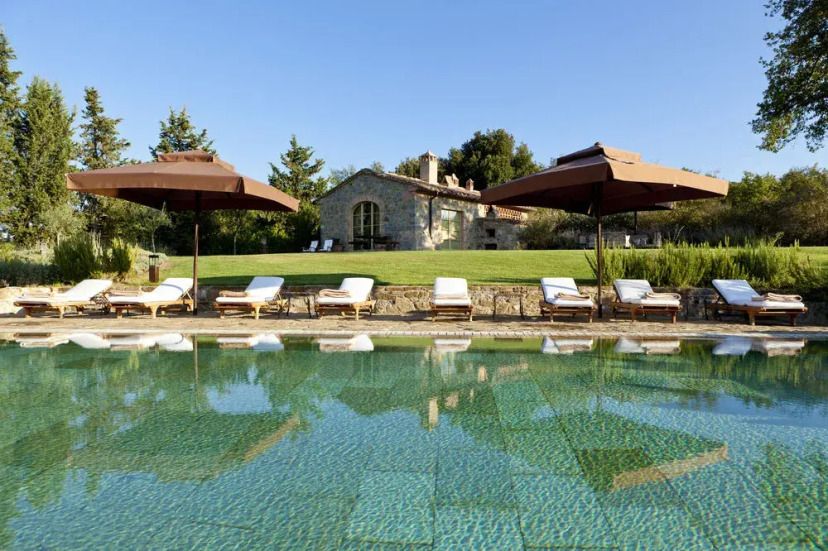 Luxuriöser Poolbereich im Rosewood Castiglion del Bosco mit Sonnenliegen und großen Sonnenschirmen, vor einer charmanten Steinvilla inmitten der toskanischen Hügellandschaft unter klarem Himmel.