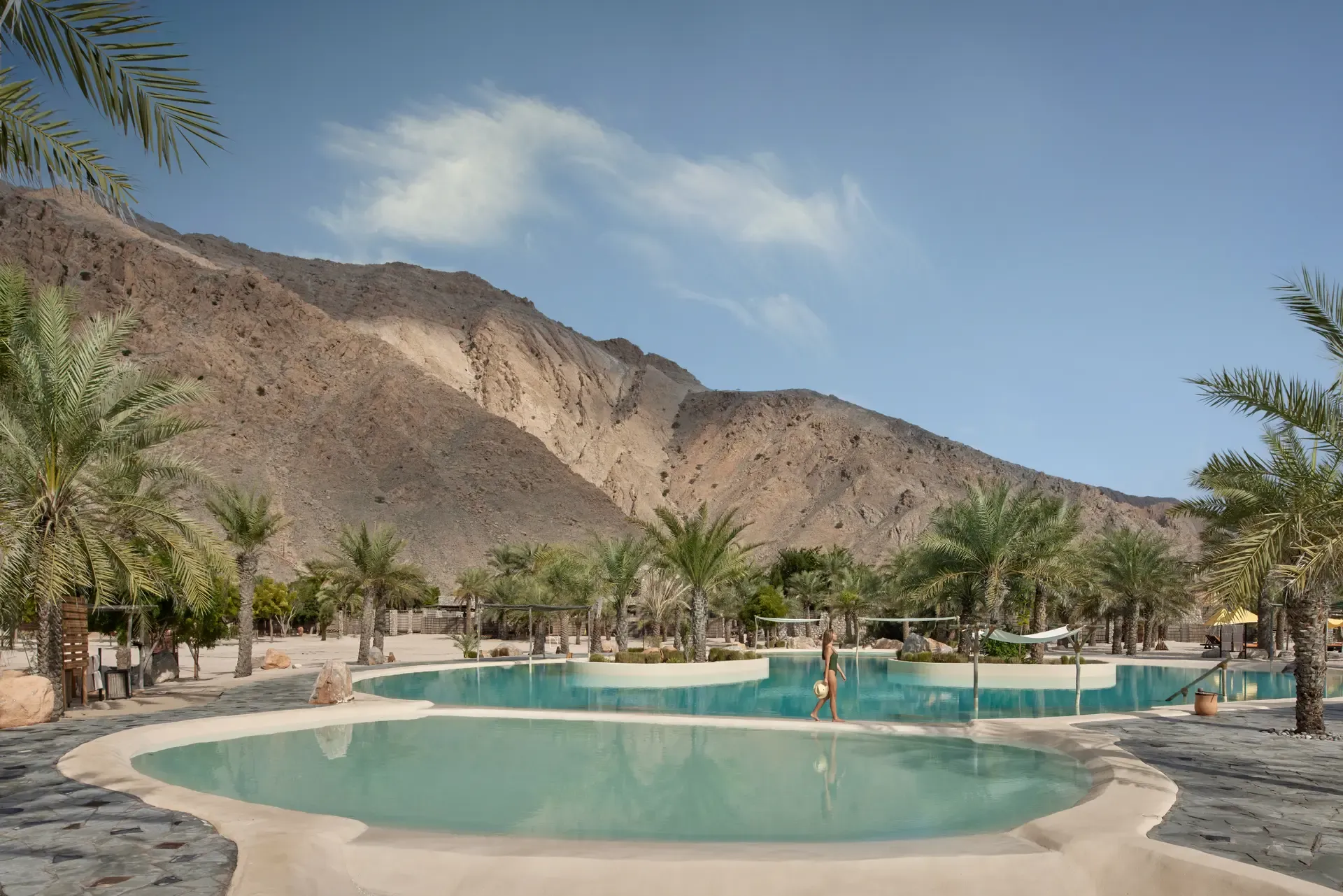 Blick auf den palmengesäumten Pool des Six Senses Zighy Bay Resorts im Oman, umgeben von einer majestätischen Felskulisse und Wüstenlandschaft, mit einer Person, die entspannt am Wasser entlanggeht.